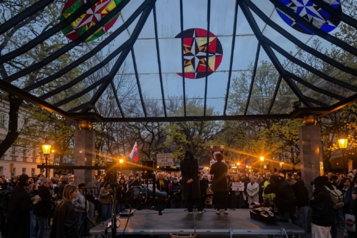 Crowd standing around two people on elevated stage, streetlights casting light. 