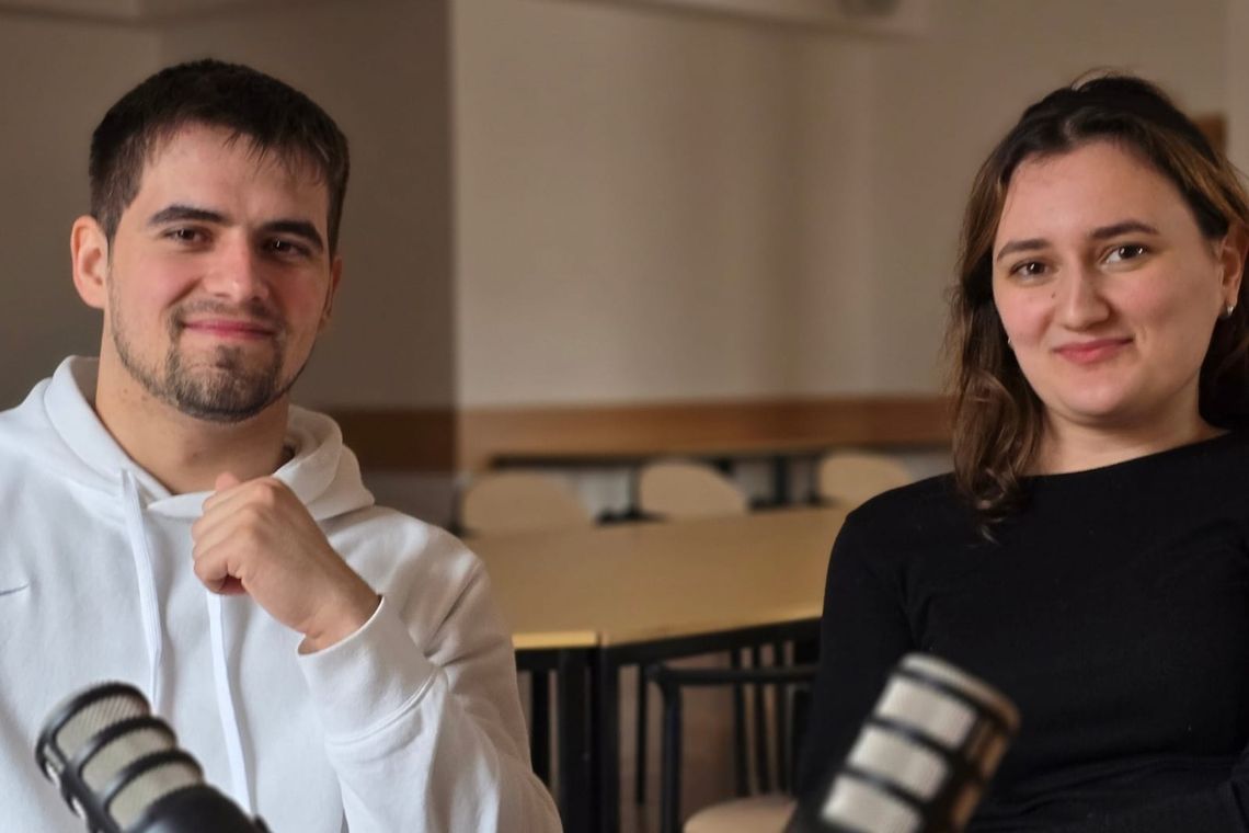 Two people looking at camera with microphones in front of them and table and chairs in background. 