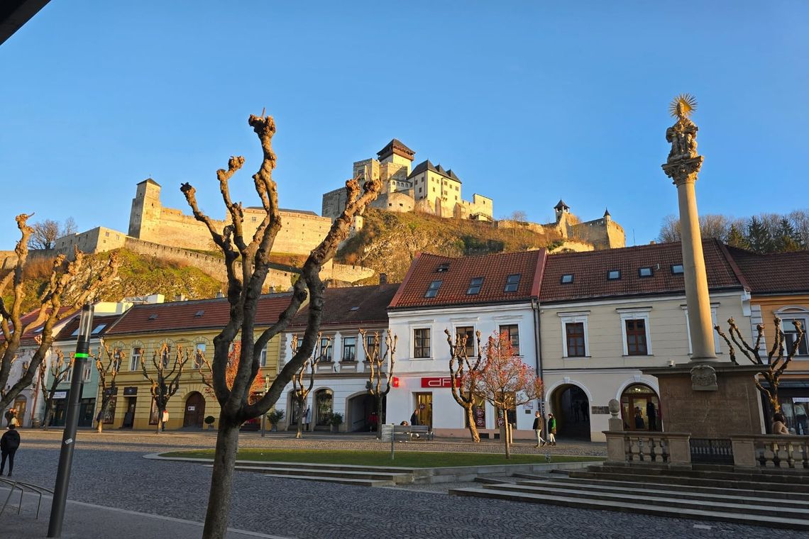 Buildings in a row with a monument and some bare trees in foreground and a castle on a hill in background. 