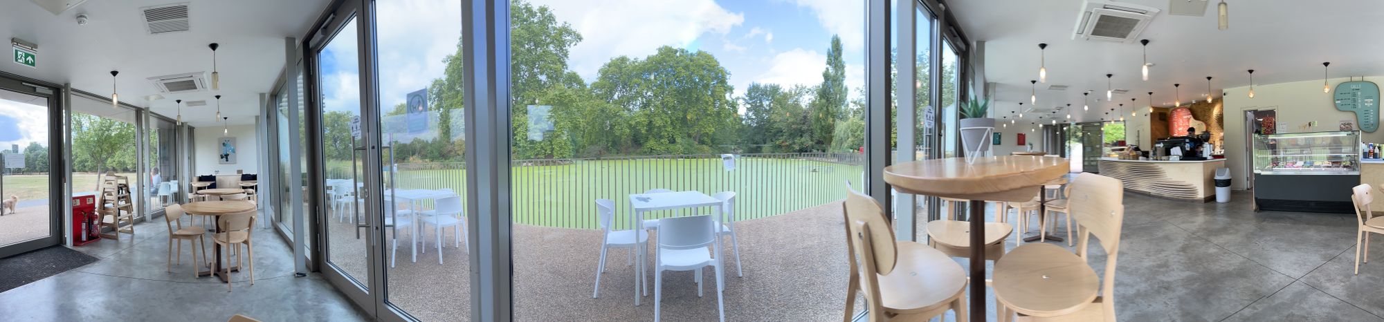 Panorama of a cafe, with glass windows to a lake covered in green algea with trees behind. To the left side is more cafe, wit