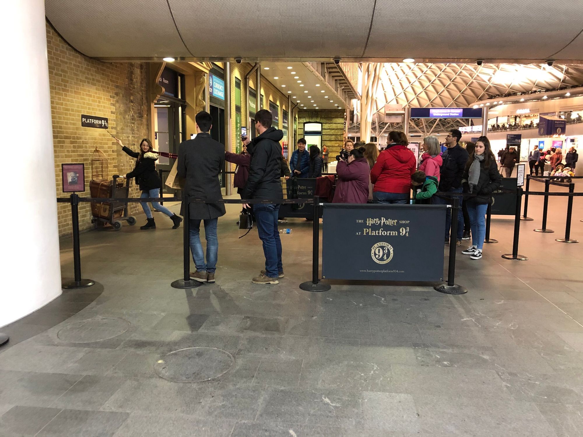Half a luggage trolly is up against a brick wall as if disappearing into it. A woman poses pushing the trolley. Behind her is