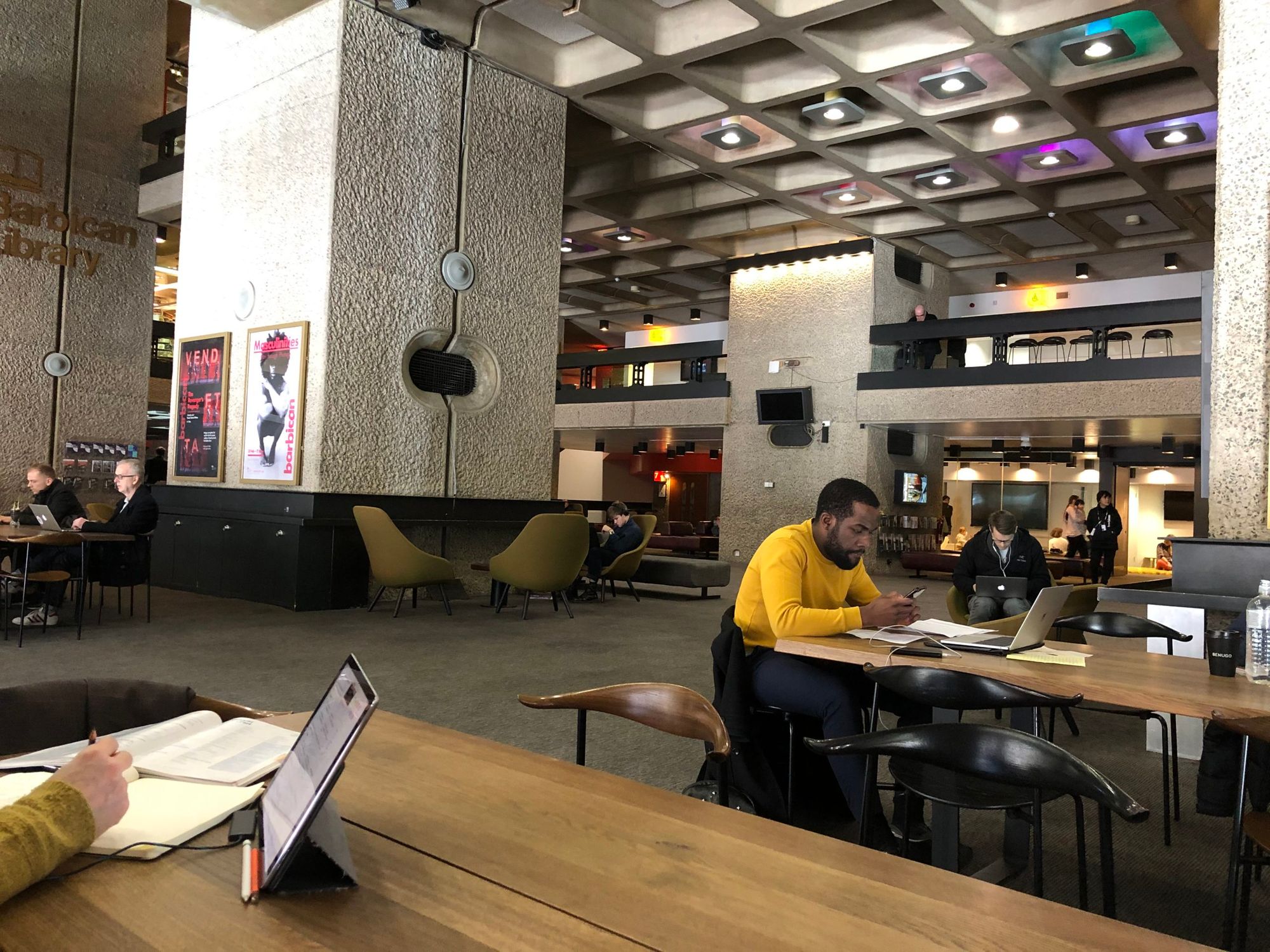 A large boxy granate hall, people work on laptops and drink coffee in the hall and on a mezzanine balcony above. Stylish furn