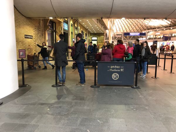 Half a luggage trolly is up against a brick wall as if disappearing into it. A woman poses pushing the trolley. Behind her is