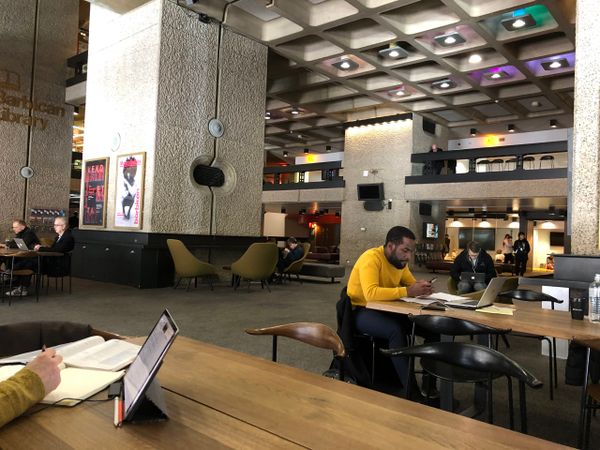 A large boxy granate hall, people work on laptops and drink coffee in the hall and on a mezzanine balcony above. Stylish furn