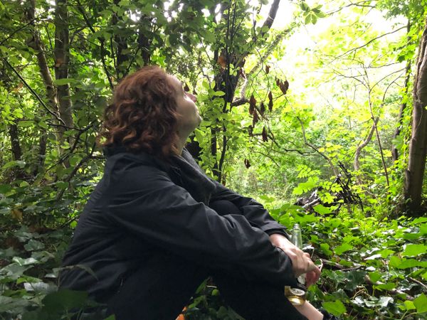 Woman sat in the woods looks up wistfully. She holds a bottle of beer. The remains of a rusted moped can be seen in the backg