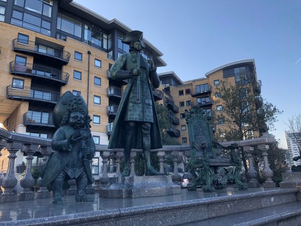 Statue of Peter The Great standing on a plinth. To the left is a statue of a court dwarf and to the right is a statue of a th
