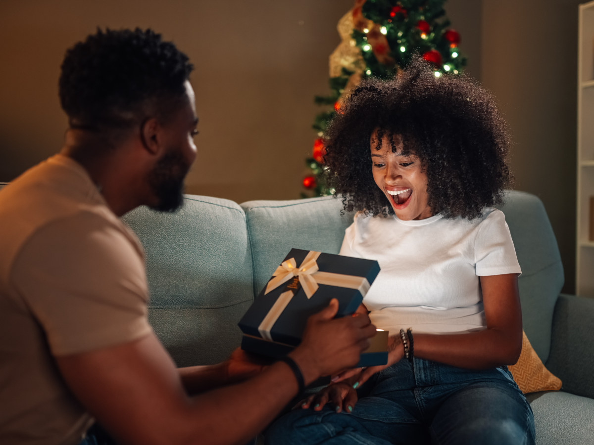 Person giving a wrapped present to their partner, who looks surprised and delighted while sitting near a decorated Christmas tree.