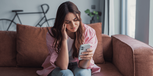Woman sitting on a couch holding a phone, touching her temple with a worried expression in a living room with a bicycle in the background.