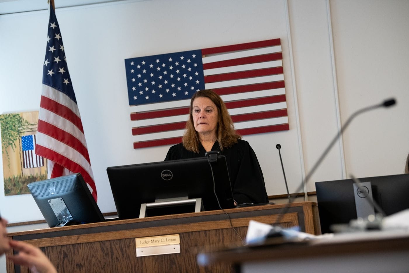 A white woman with brown hair standing at a judge's bench looking out over the courtroom. She is wearing black judges robes.