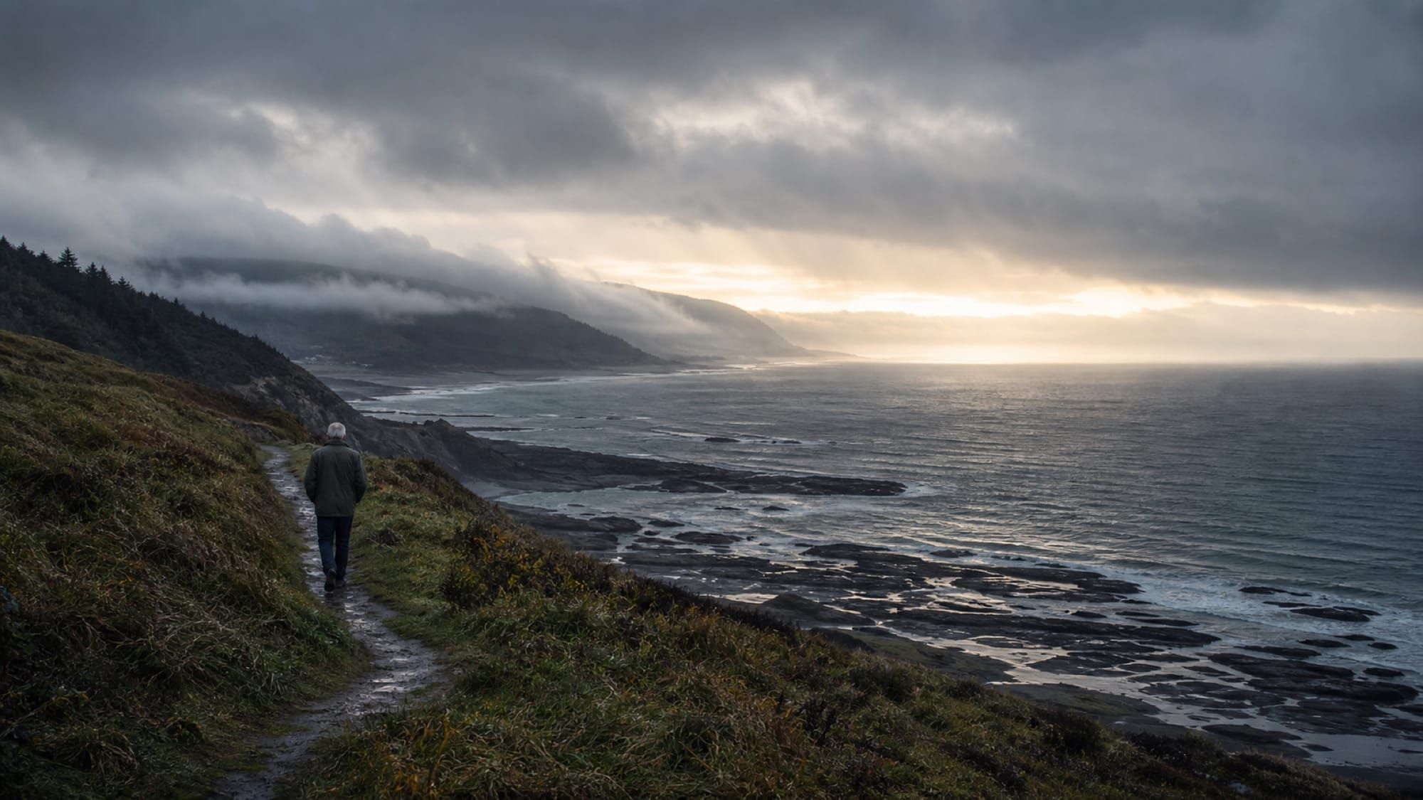 A lone figure walks a coastal path under a wide, overcast sky at dusk.