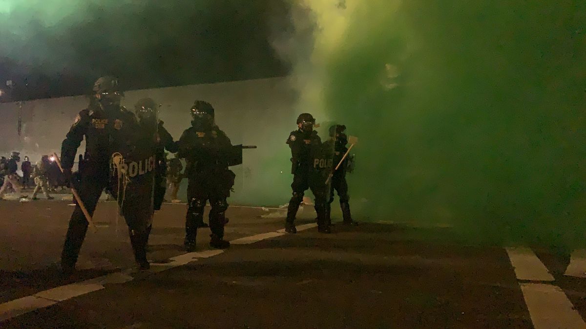 Photo of DHS police holding a line outside of Portland ICE. They are carrying shields, bats, and there is a vibrant thick green cloud of smoke behind them from munitions the feds deployed