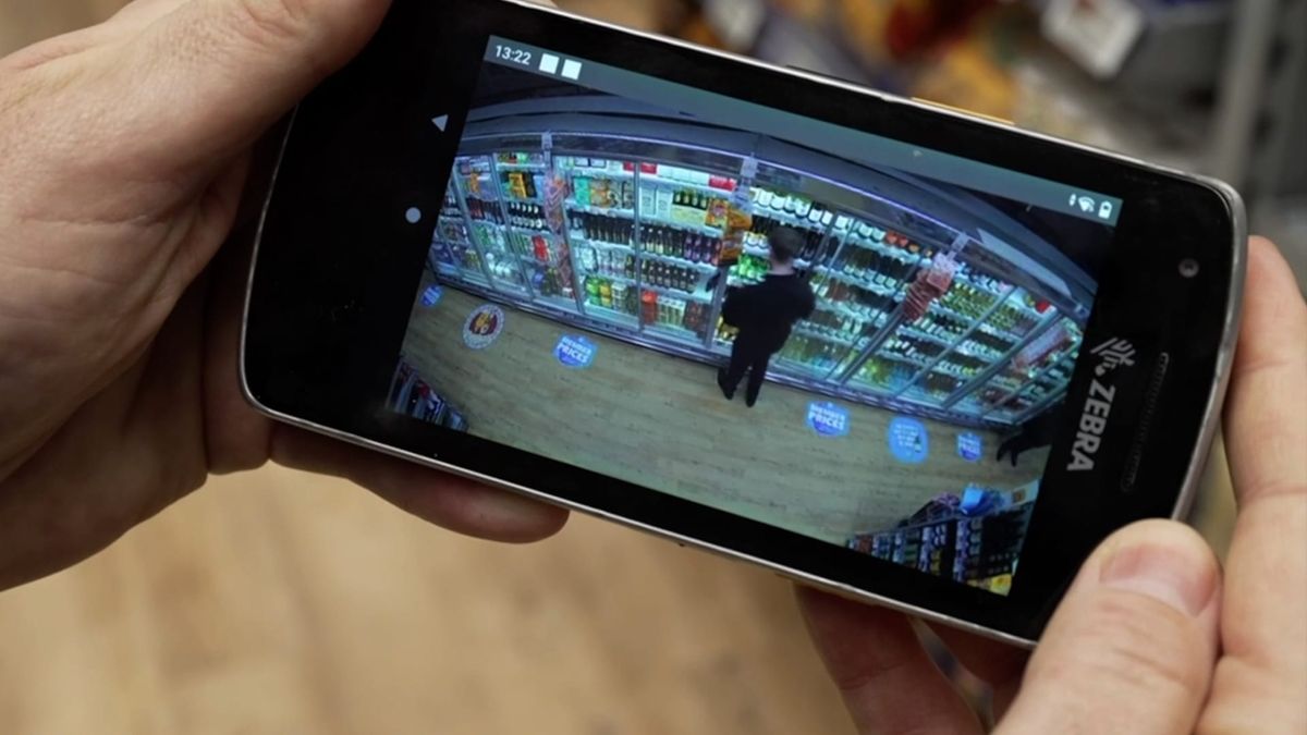 Hands holding a smart-phone type device showing an image of a shoplifter taking items from a wine fridge in a supermarket