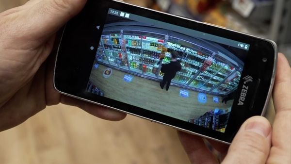 Hands holding a smart-phone type device showing an image of a shoplifter taking items from a wine fridge in a supermarket