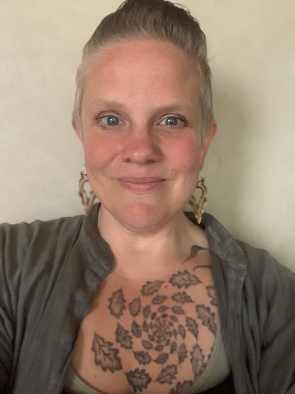 AG Derwen, wry smile, white bodied, femme presenting, short hair, oak leaf tattoo over heart