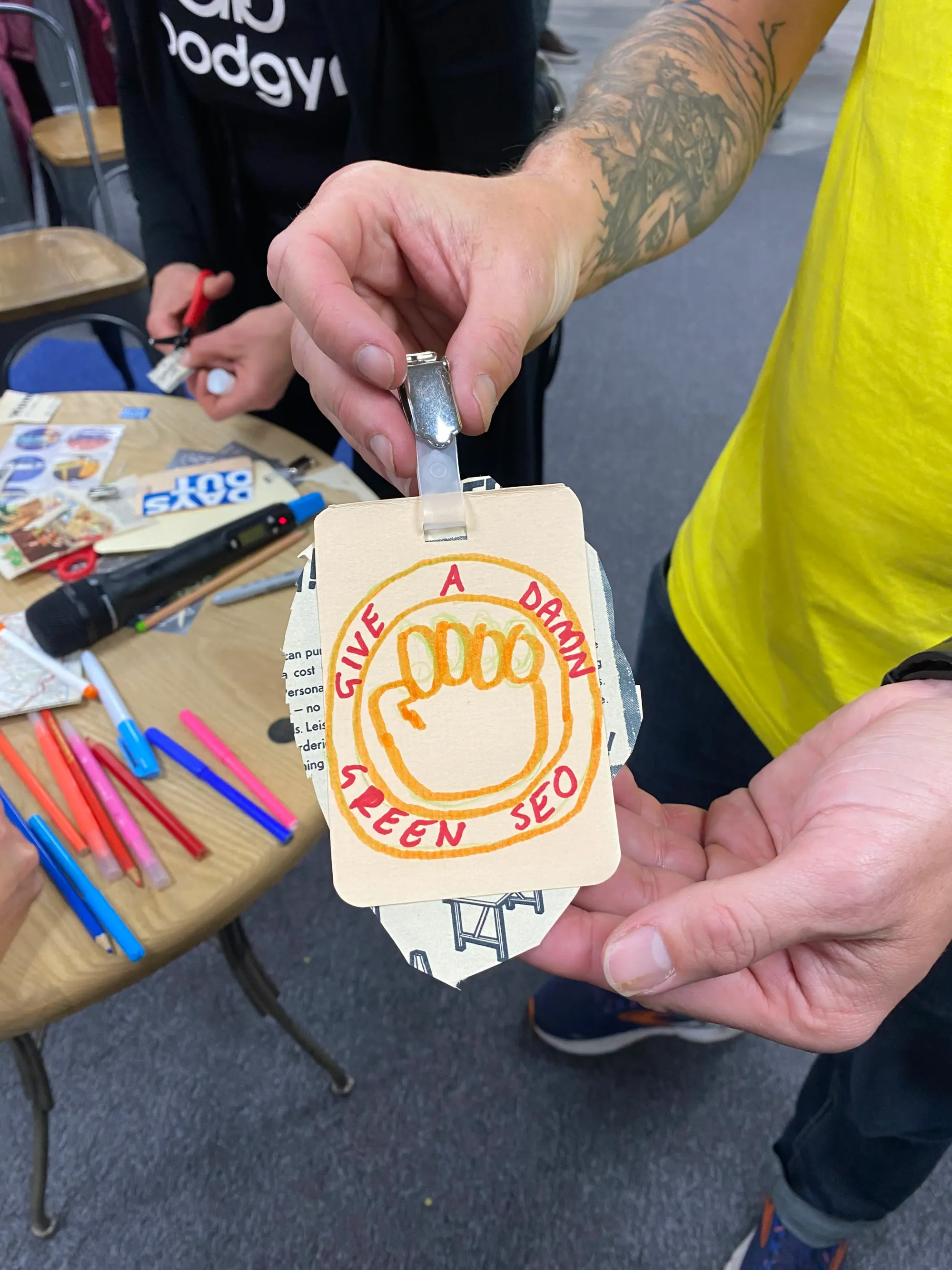 Close-up of hands holding a hand-drawn protest badge. The text on the badge says "Give a Damn. Green SEO"