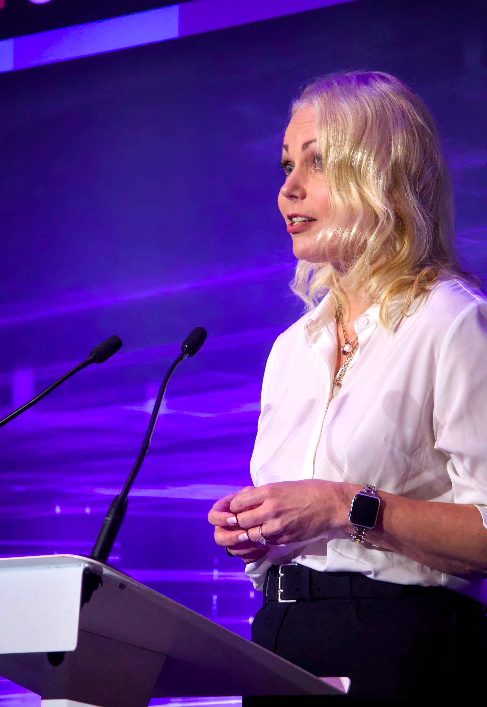 A woman with long blonde hair, wearing a white blouse and black trousers, stands at a podium with two microphones. She holds her hands together while speaking, wearing a smart watch and necklace. The backdrop glows with purple and blue lighting, suggesting a conference or keynote presentation.