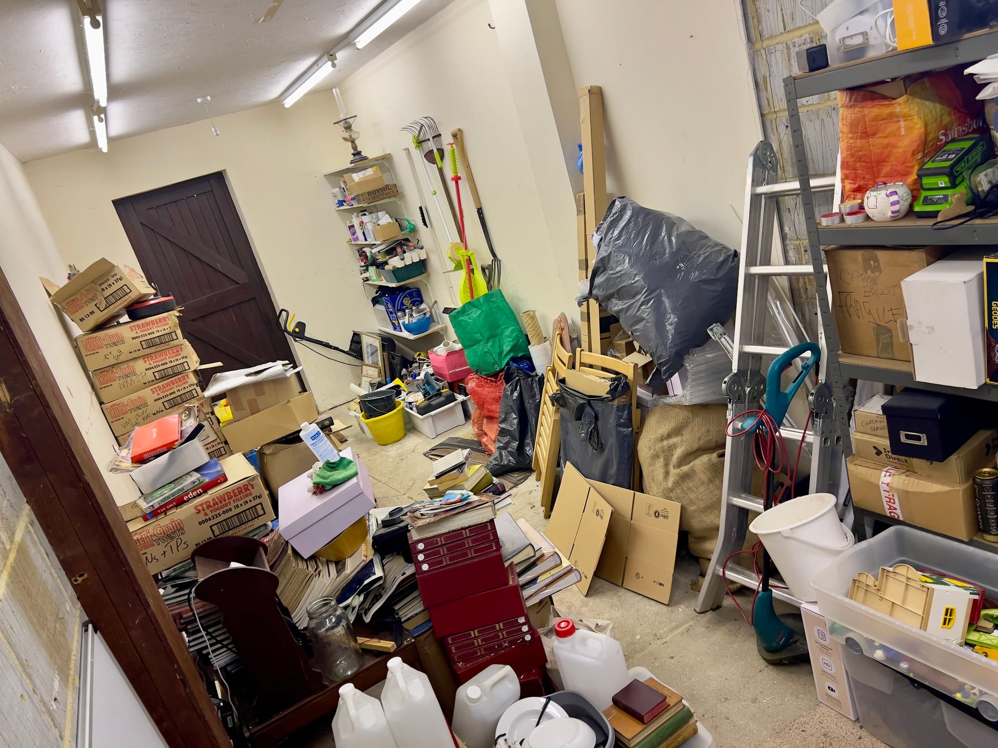 Cluttered garage or storage room filled with stacked boxes, tools, shelves, cleaning supplies, books and household items, with a ladder leaning against shelving on the right.