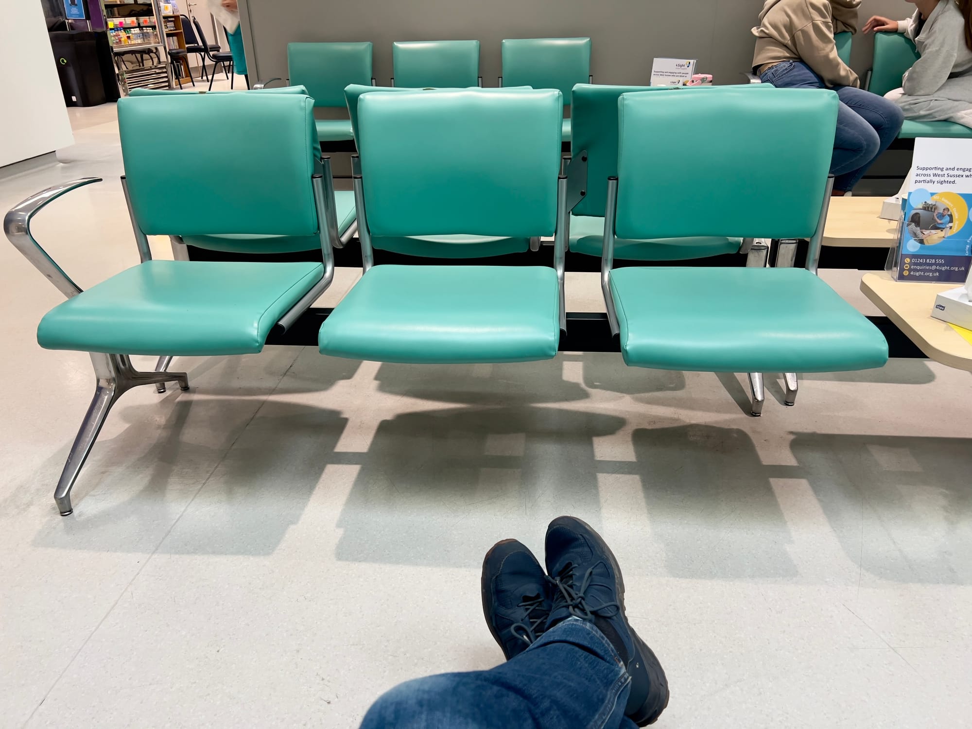 Rows of empty turquoise chairs in a hospital or clinic waiting room, viewed from a seated person’s perspective with crossed legs and dark trainers visible in the foreground.