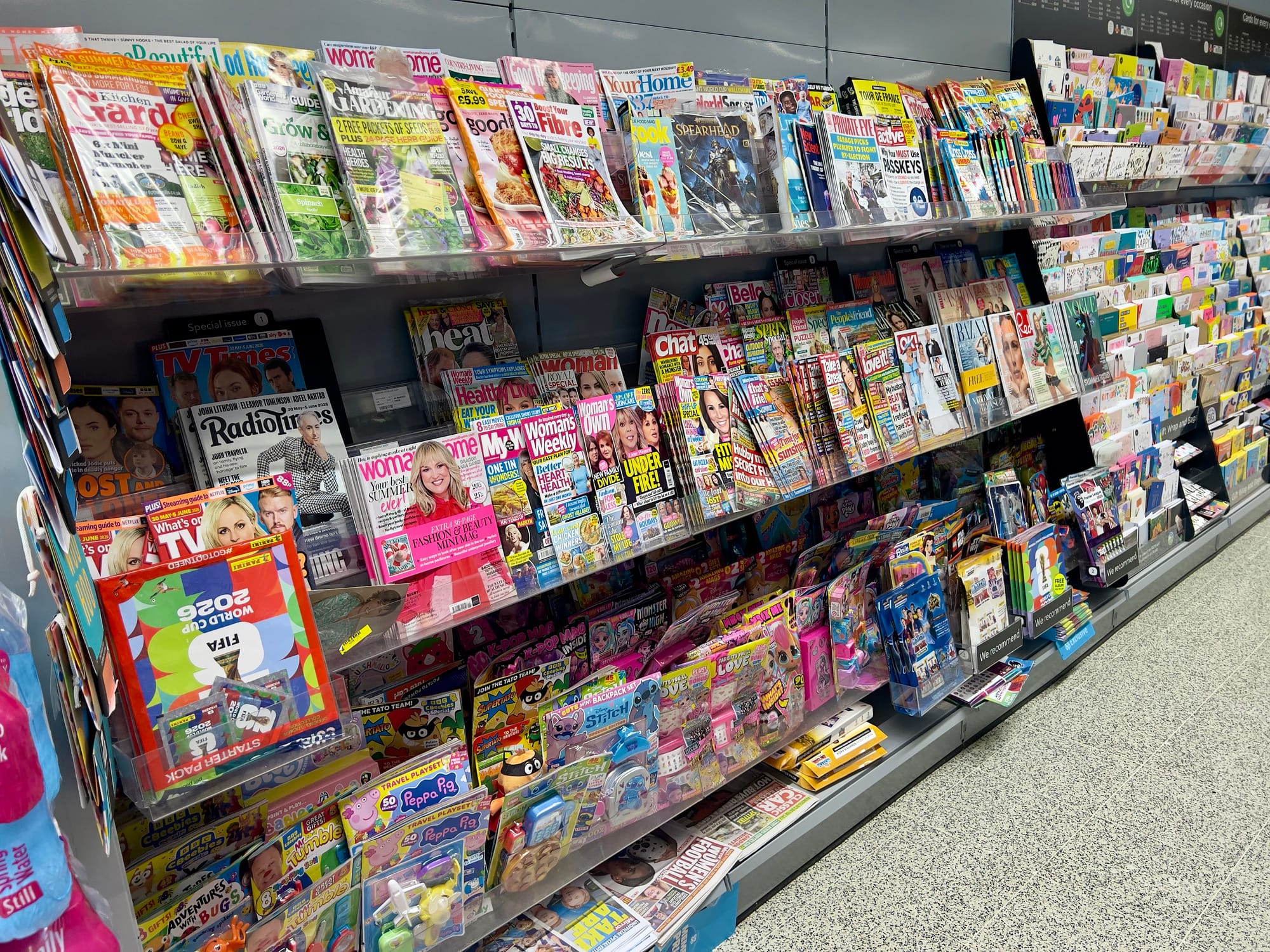 Magazine and greeting card display in a supermarket or newsagent, with shelves packed with colourful magazines, puzzle books, children’s activity packs and cards stretching along a wide aisle.