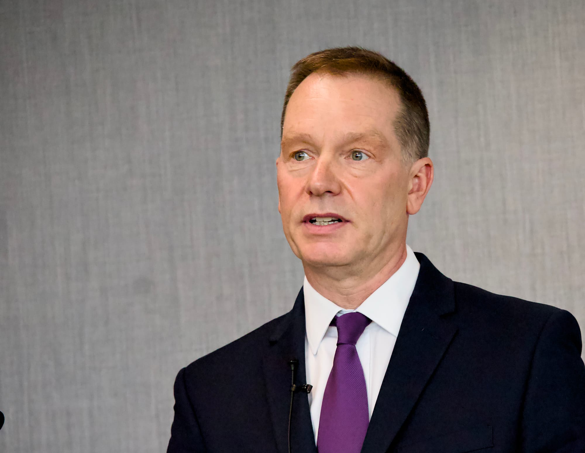 A middle-aged man with short grey-brown hair and light eyes, wearing a dark navy suit jacket, white dress shirt, and purple tie, speaks at an event. A small lapel microphone is visible on his lapel. The background is a plain grey textured surface.