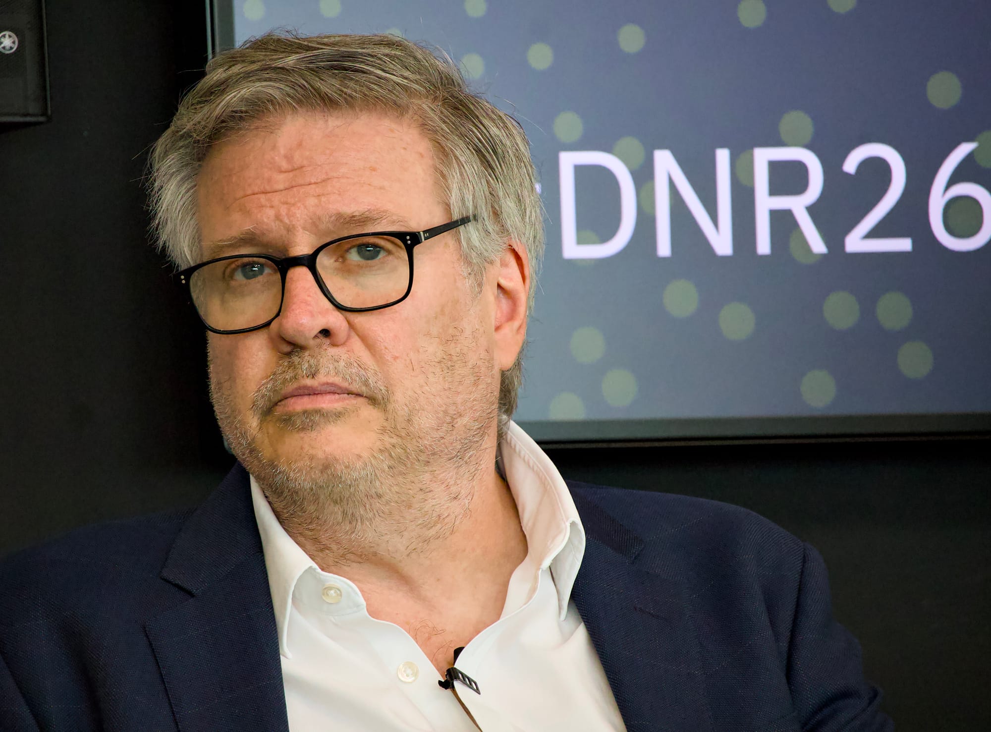A middle-aged man with grey hair, short stubble, and black rectangular-framed glasses, wearing a navy blazer over an open-collar white shirt, with a lapel microphone clipped to his collar. He is seated and gazing forward with a thoughtful expression. Behind him is a dark screen displaying the partial text 'DNR26' with a green polka dot pattern.