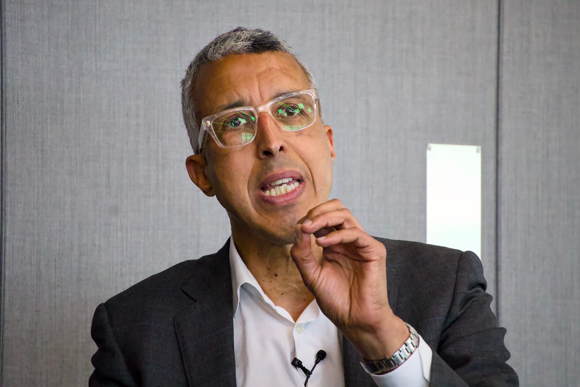 Kamal Ahmed, representing Fortune, speaking at a panel event. He has short salt-and-pepper hair and is wearing clear-framed glasses with green reflections visible in the lenses, a dark grey suit jacket, and an open-collar white shirt with a lapel microphone. He holds one hand raised to his chin in a thoughtful gesture. The background is a plain grey textured wall