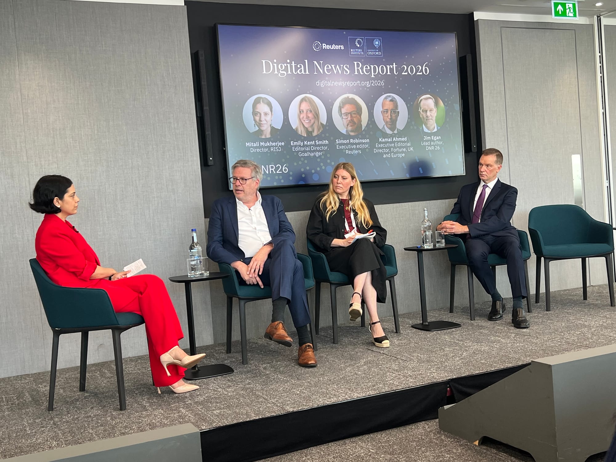 Four panellists seated in teal chairs on a raised stage at the launch event for the Reuters Institute Digital News Report 2026. From left to right: Mitali Mukherjee, Director of RISJ, wearing a red trouser suit; Simon Robinson, Executive Editor at Reuters, in a navy blazer and open-collar white shirt; Emily Kent Smith, Editorial Director of Goalhanger, in a black jacket and dark red top; and Jim Egan, Lead Author of DNR 26, in a dark navy suit with a purple tie. Small side tables with water bottles are placed between the panellists. Behind them, a large screen displays the title 'Digital News Report 2026' alongside the Reuters Institute and University of Oxford logos, and headshots and titles of all five panellists including Kamal Ahmed, Executive Editorial Director of Fortune UK and Europe.