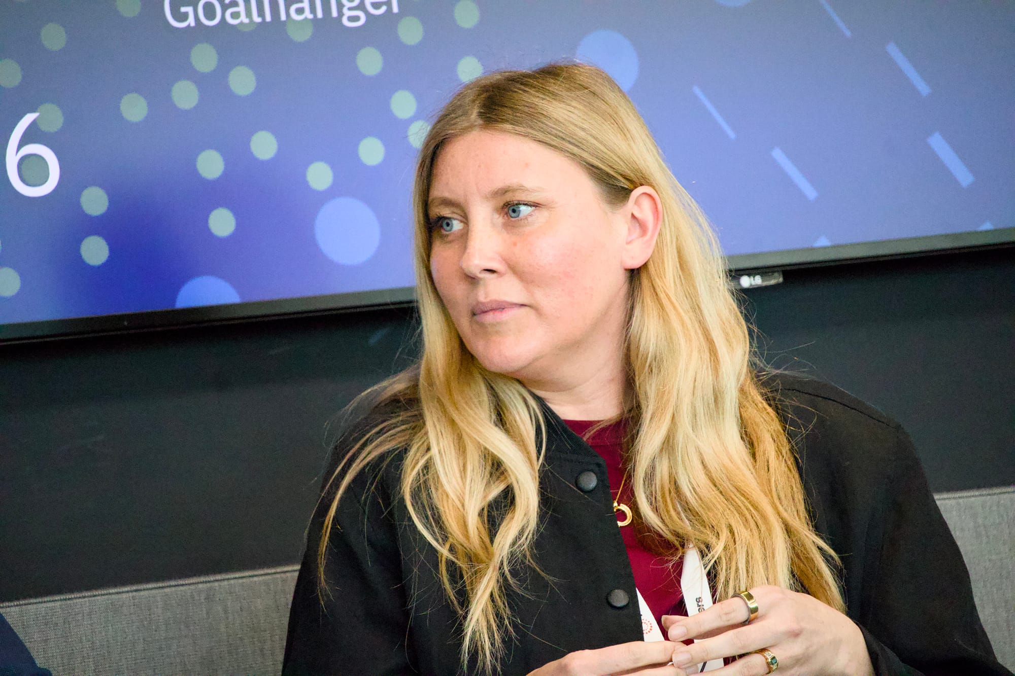 Emily Kent Smith, representing Goalhanger, seated at a panel event. She has long blonde hair and blue eyes, and is wearing a black jacket over a dark red top, with a gold necklace. She holds an event lanyard in her hands and gazes to one side with an attentive expression. Behind her is a screen displaying the word 'Goalhanger' alongside a blue and green polka dot pattern.