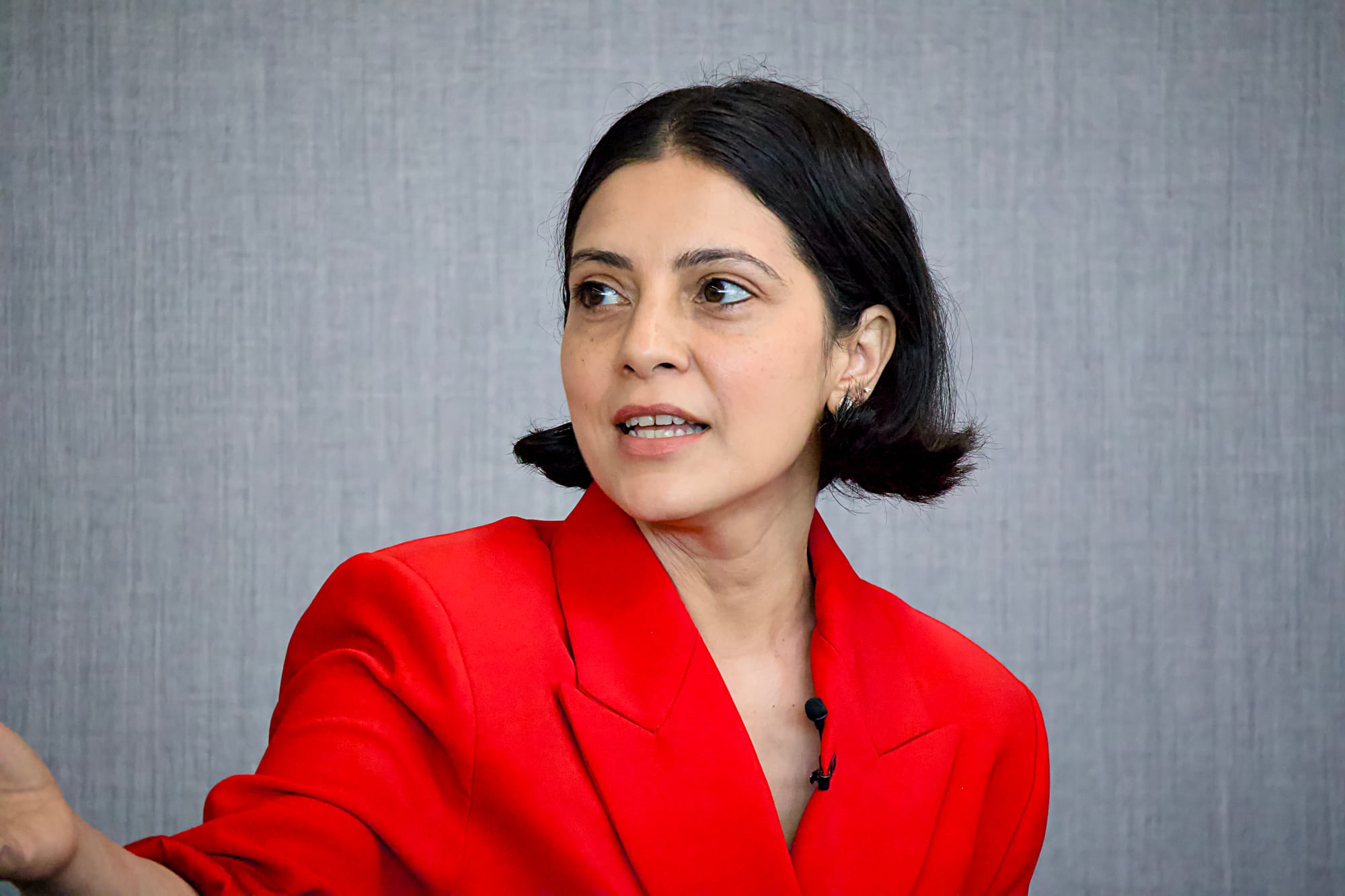 "A woman with short dark hair and dark eyes, wearing a bold red blazer, speaks at an event while glancing to one side. She has small stud earrings and a lapel microphone clipped to her jacket. The background is a plain grey textured surface.