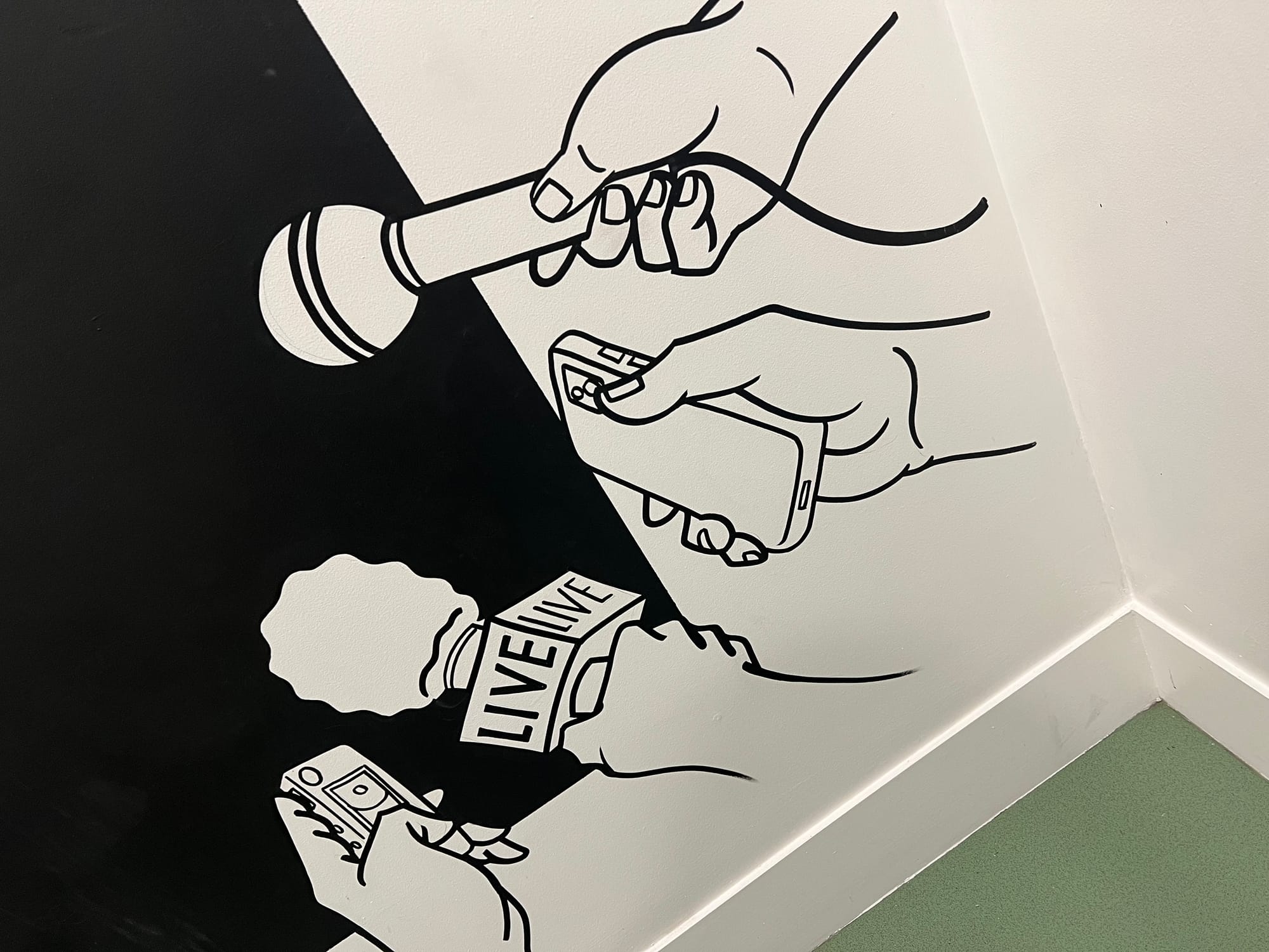 Black-and-white line-art mural painted across an interior corner wall, depicting hands holding microphones (one labeled 'LIVE') and smartphones in a repeating pattern, suggesting themes of media, journalism, and interviews.