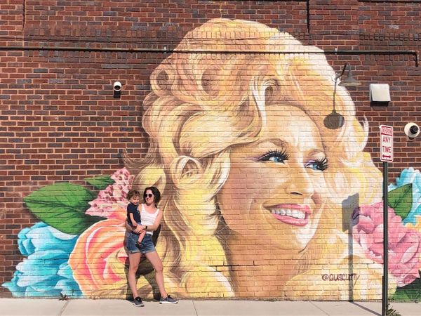 The sexiest woman alive stands in front of the Dolly Parton mural in West Asheville, holding the best and most adorable child who ever lived.