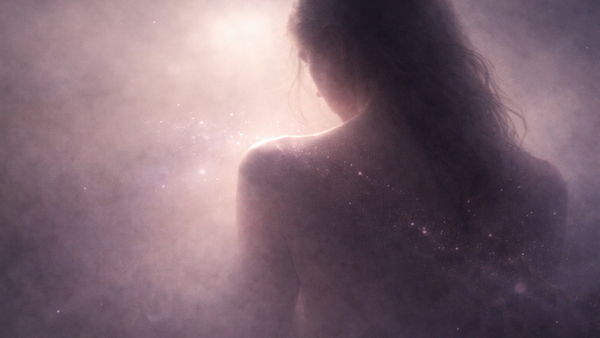 A woman is seen from behind in soft pink and violet light, with small star-like particles glowing across her back and shoulder.