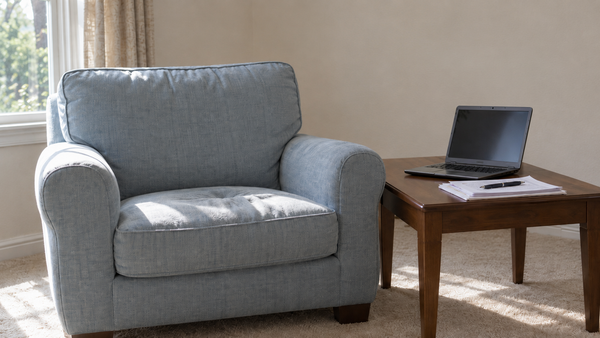 Pale blue-grey armchair by a window, its cushion still holding the hollow where someone had been sitting. Afternoon light crosses the empty seat. A laptop and papers on the side table.
