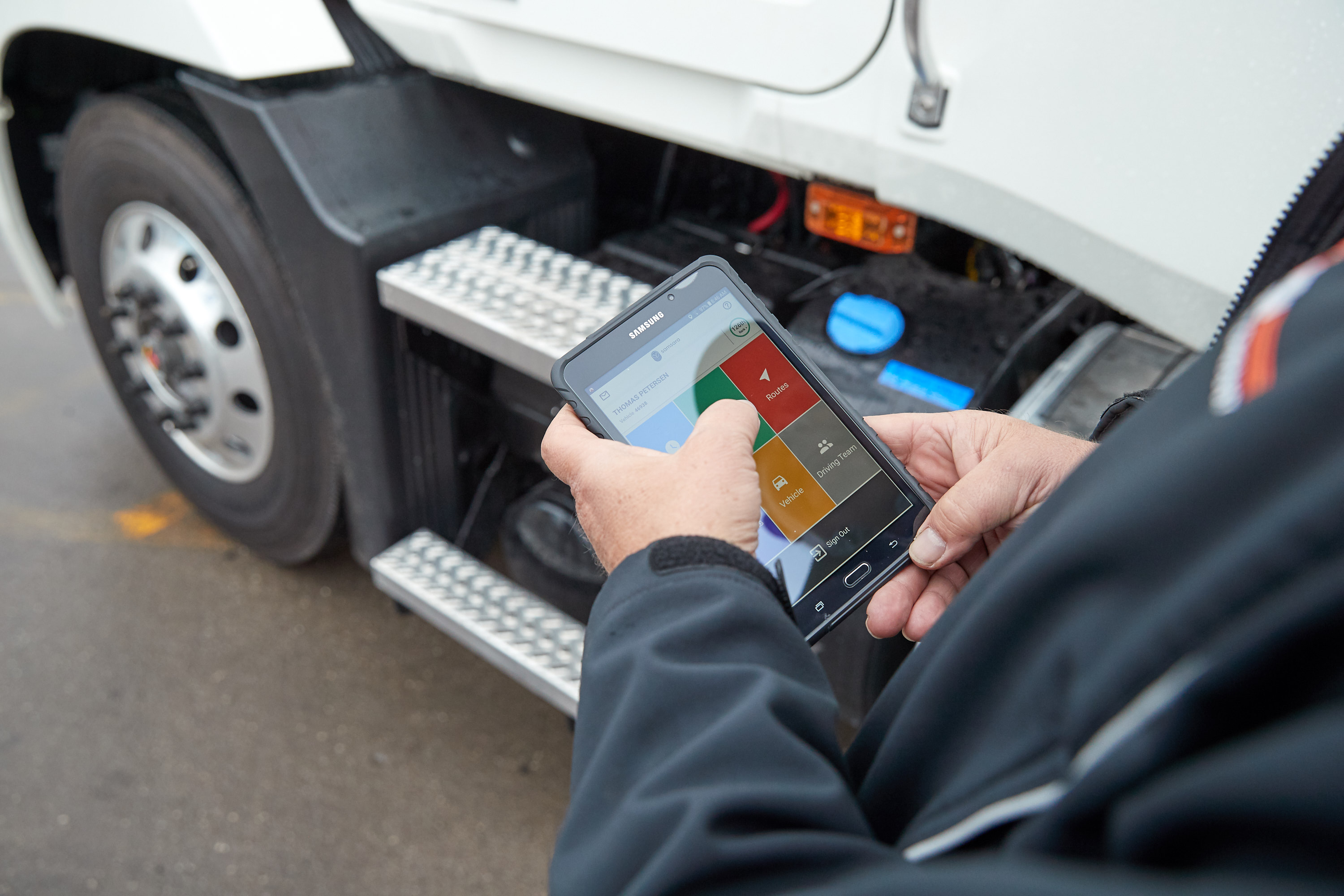 A truck driver stands beside a commercial vehicle using a Samsung tablet to access a fleet management app, showing route, vehicle, and driving data—illustrating Samsara’s mobile tools for monitoring and improving driver performance.