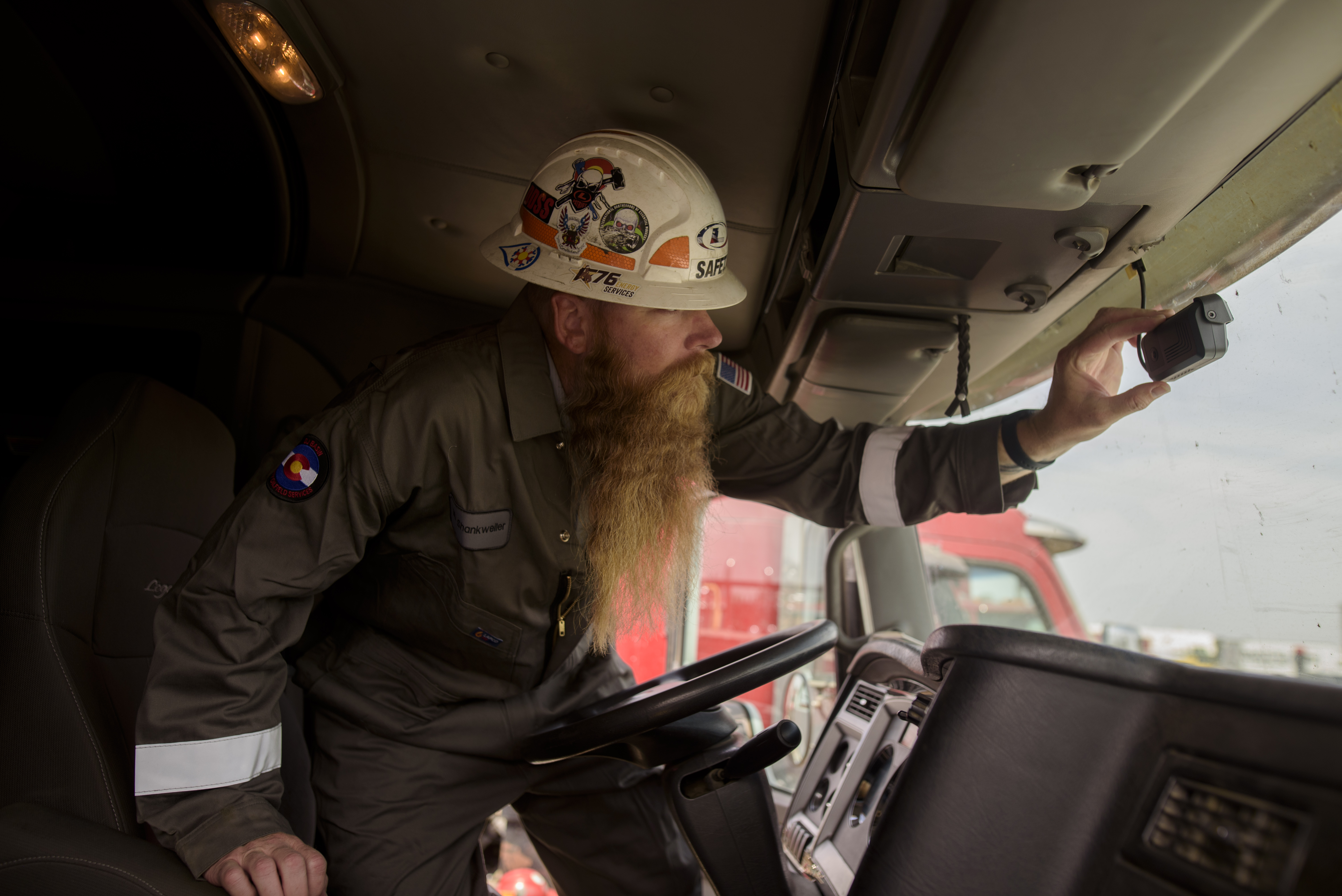 A truck driver wearing a safety helmet decorated with stickers adjusts a small dash-mounted camera inside the cab of a commercial truck, illustrating Samsara’s in-vehicle AI system used to monitor driving behavior and improve safety.