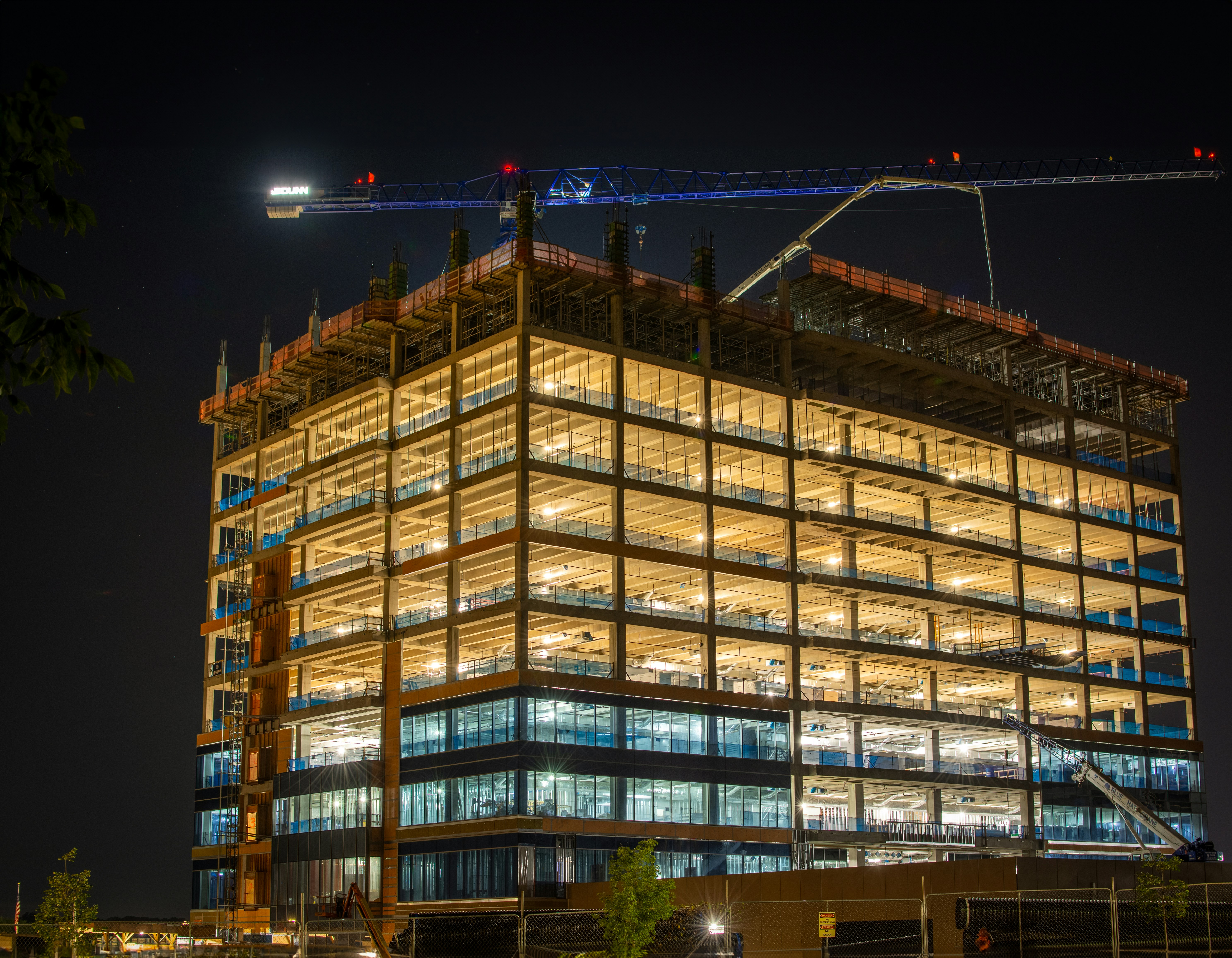 A multi-story office building under construction at night, illuminated with bright interior lights. A large crane towers above the structure against the dark sky.
