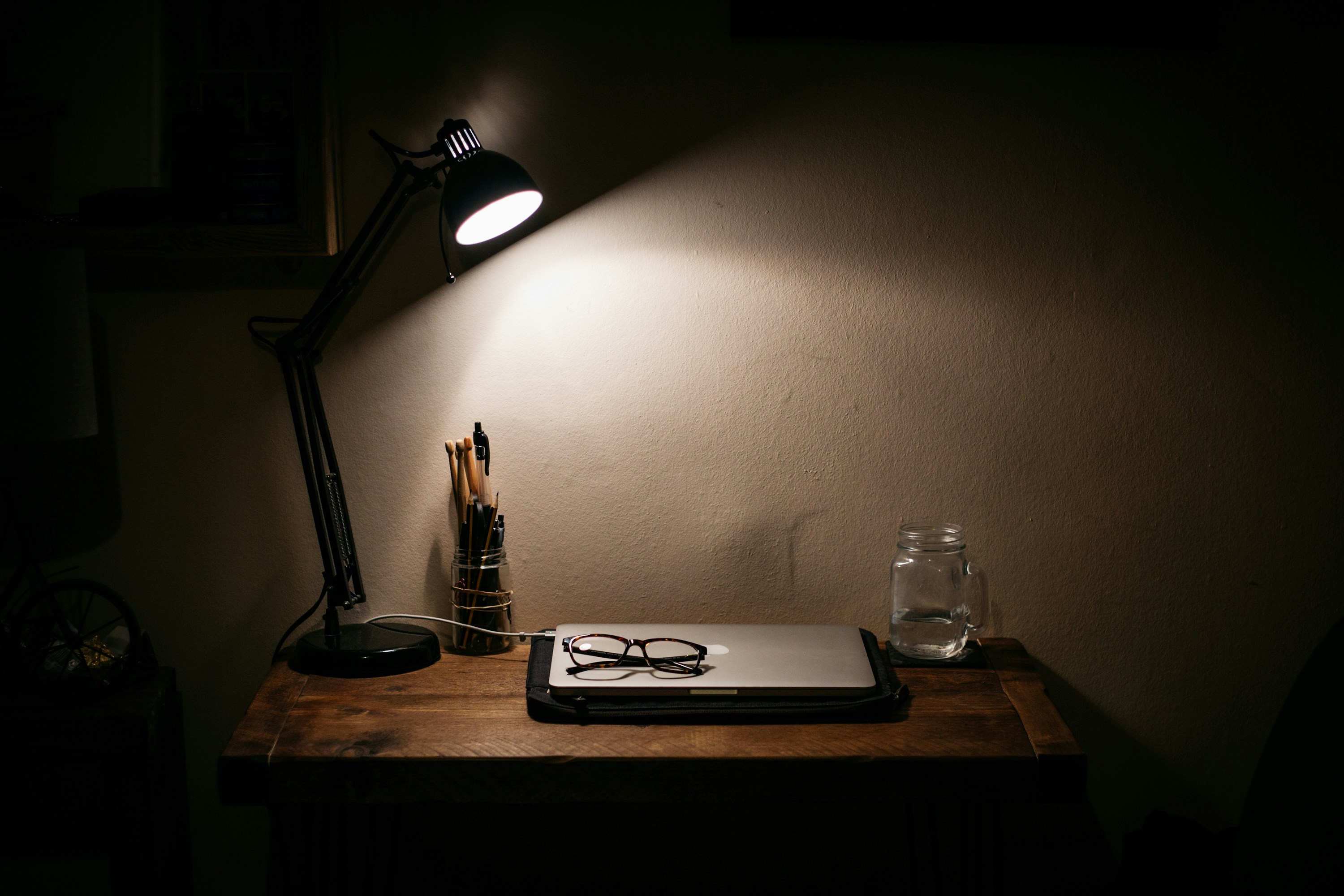 Desk with lamp on casting a shadow on a closed computer with a pair of reading glasses folded on top.