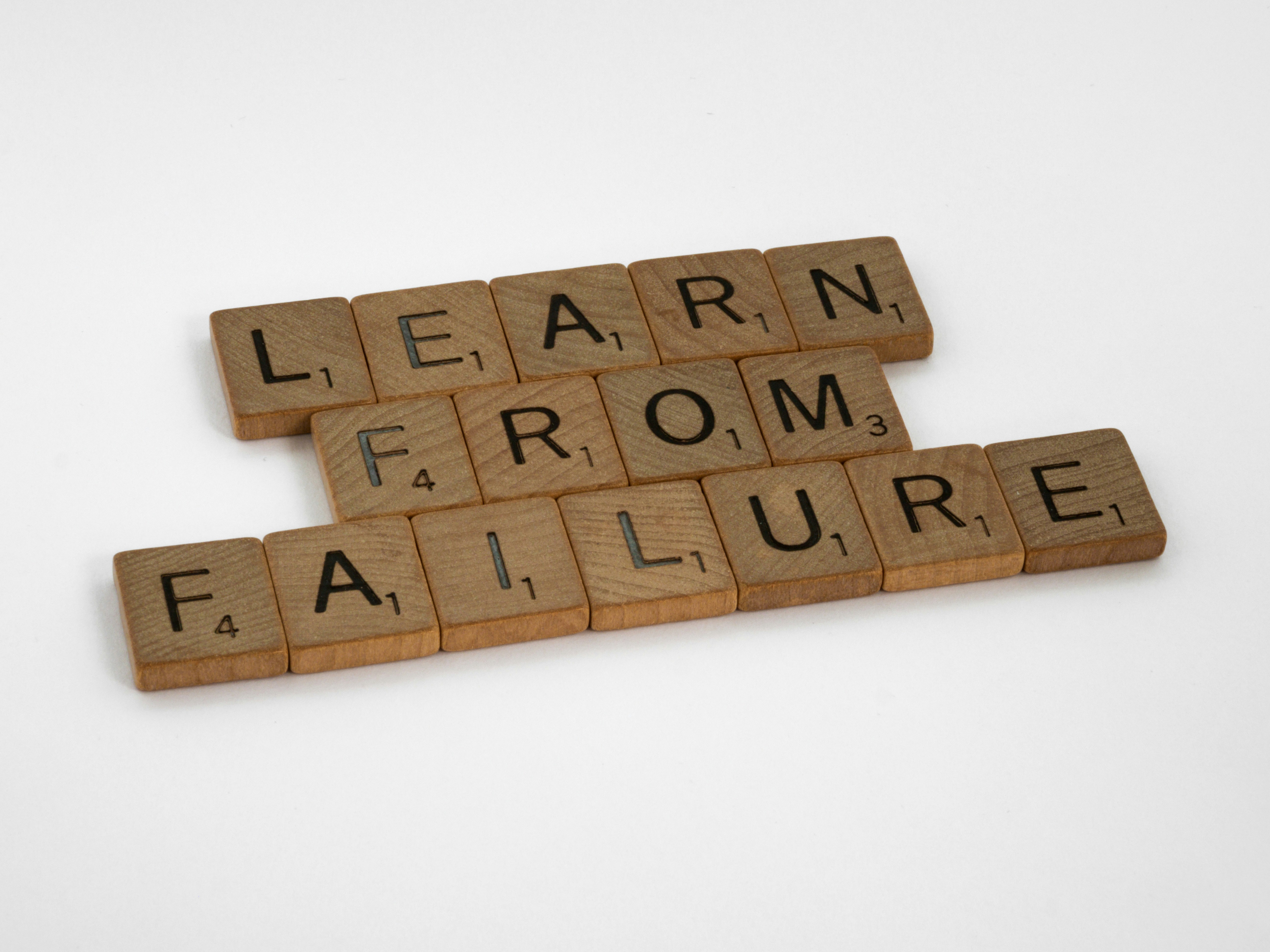 Wooden Scrabble tiles arranged on a white surface spelling the phrase “LEARN FROM FAILURE.”