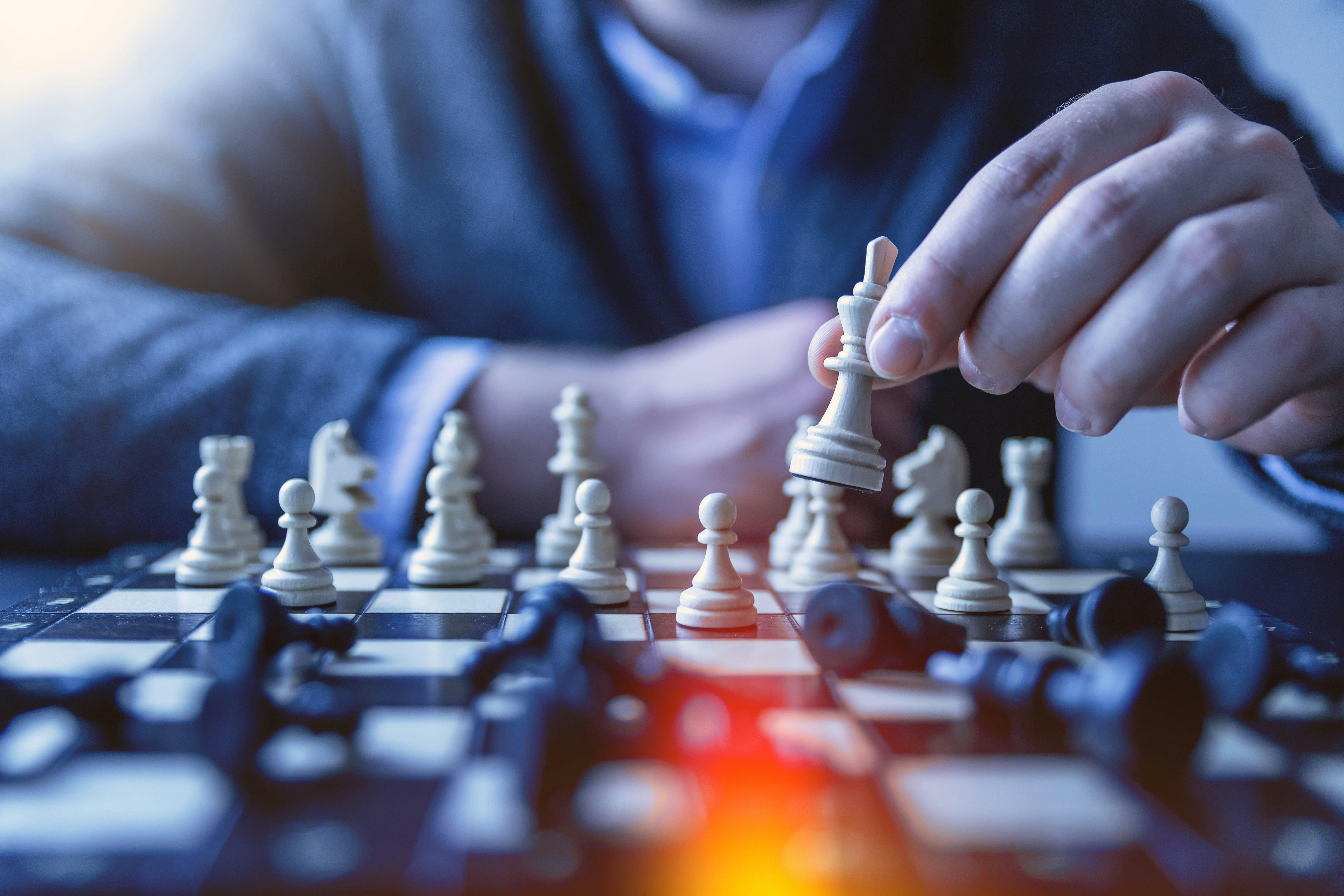 Close-up of a person playing chess, moving a white king piece on a board with scattered black and white pieces, symbolizing strategy and decision-making.