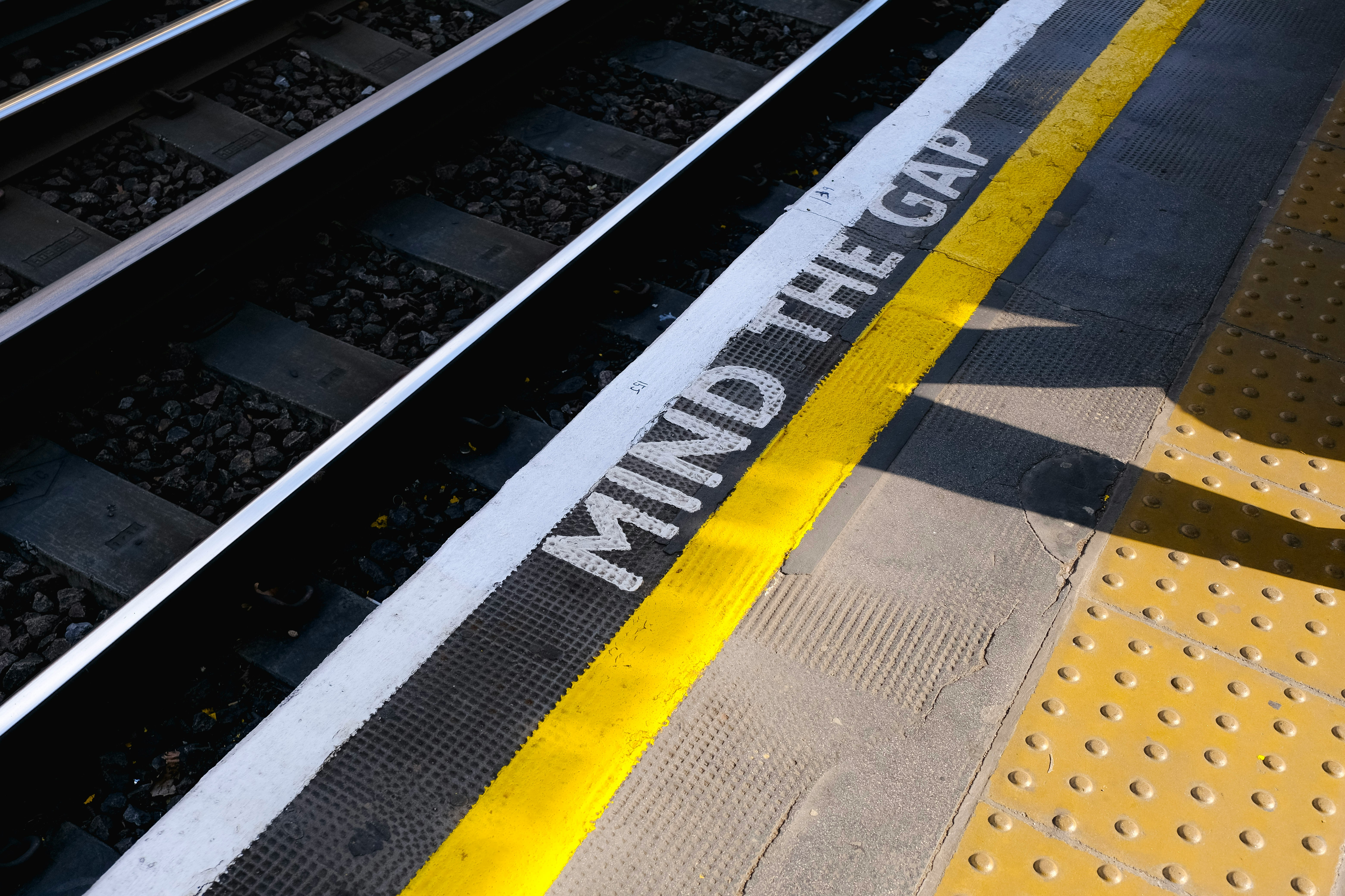 A train platform edge with a bright yellow safety line and the words “MIND THE GAP” painted beside the tracks.