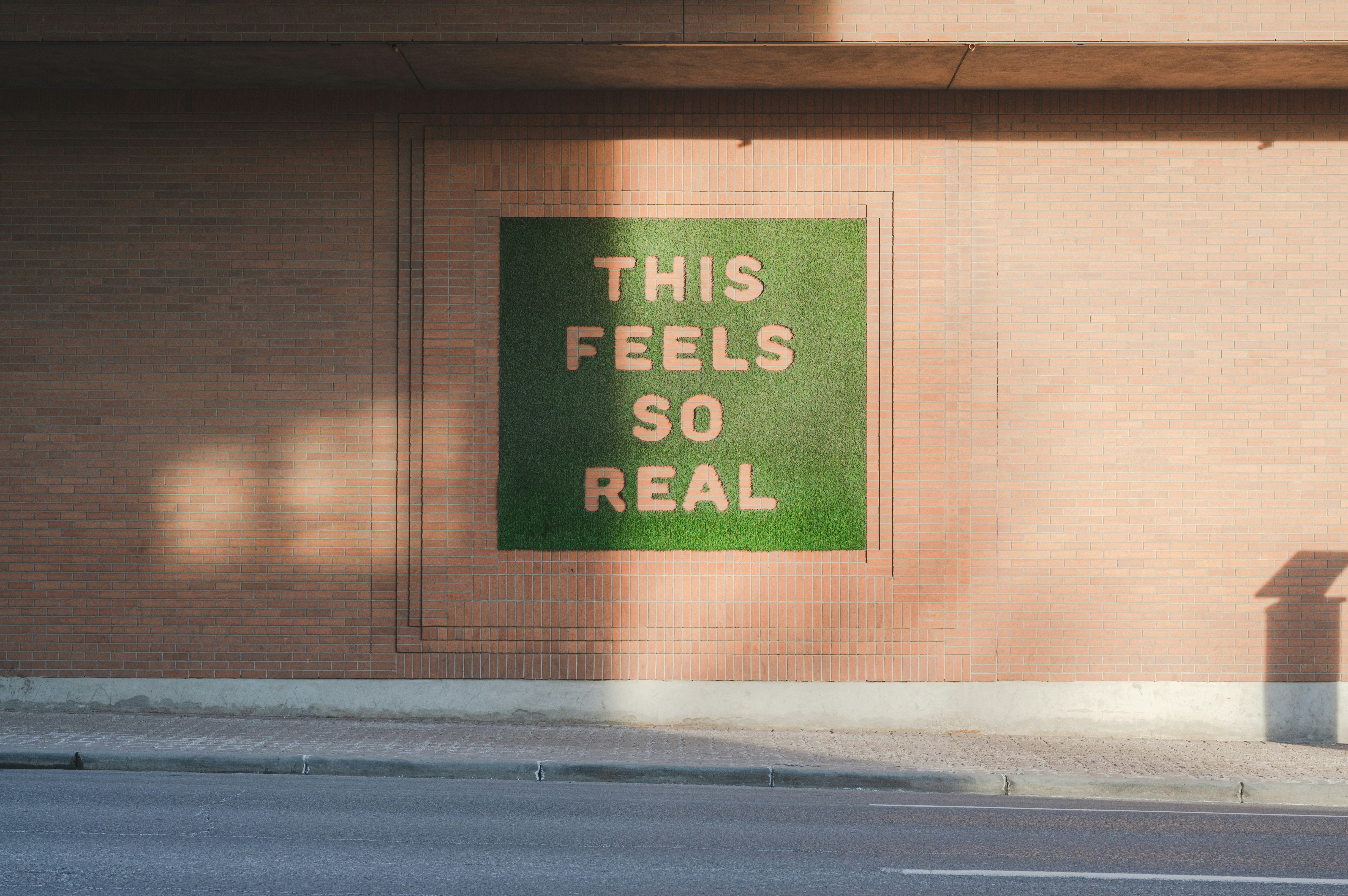 Street view of a brick wall featuring a square patch of green turf with the words “THIS FEELS SO REAL” cut out in large block letters.