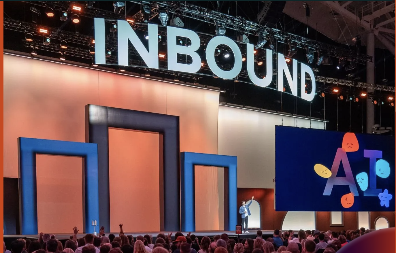 A speaker on stage at the HubSpot Inbound conference, gesturing towards a large screen with a presentation about AI, in front of a large, seated audience.