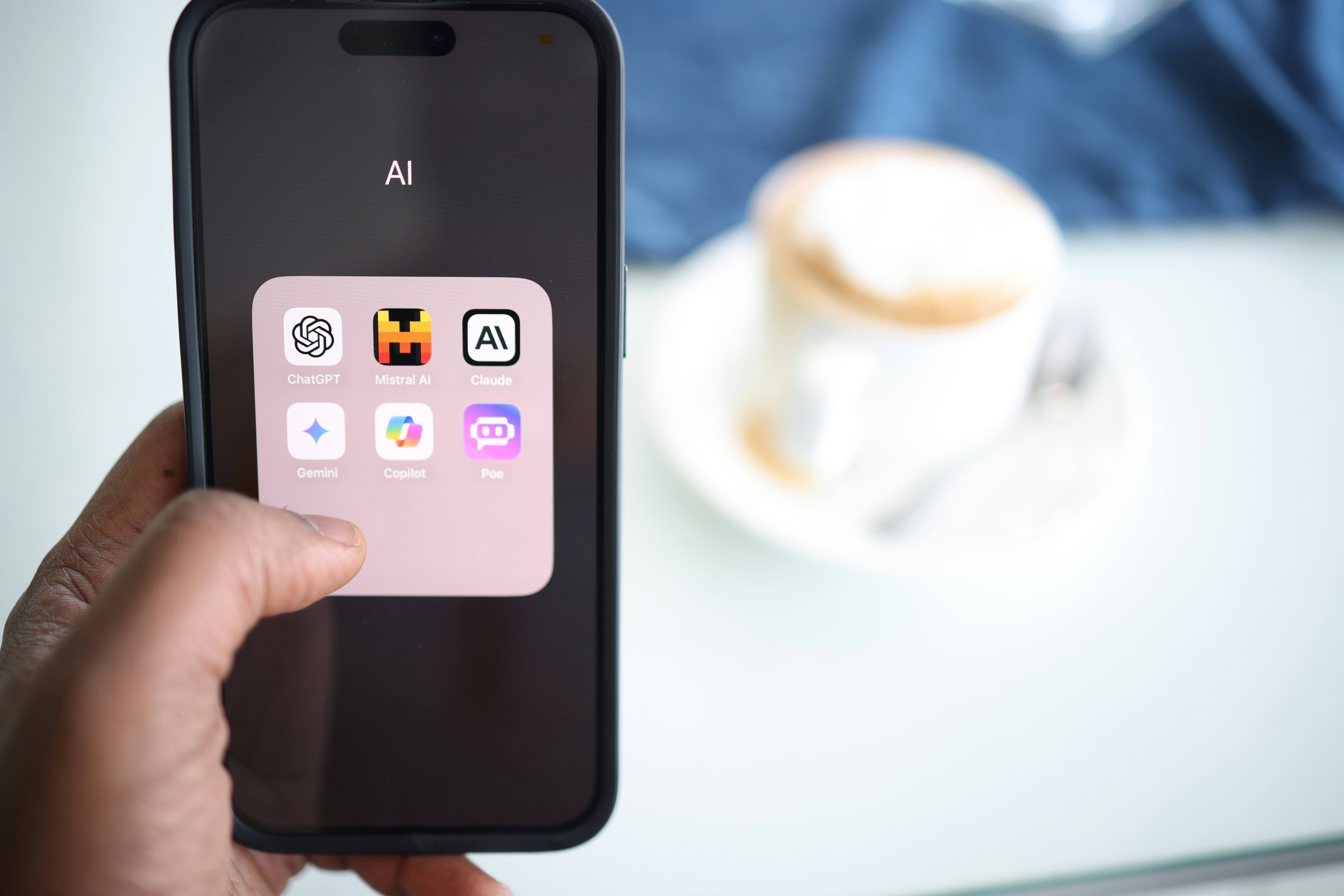 A person holds a smartphone displaying a folder labeled “AI” with icons for ChatGPT, Mistral AI, Claude, Gemini, Copilot, and Poe apps. A cup of coffee and saucer are blurred in the background on a table.