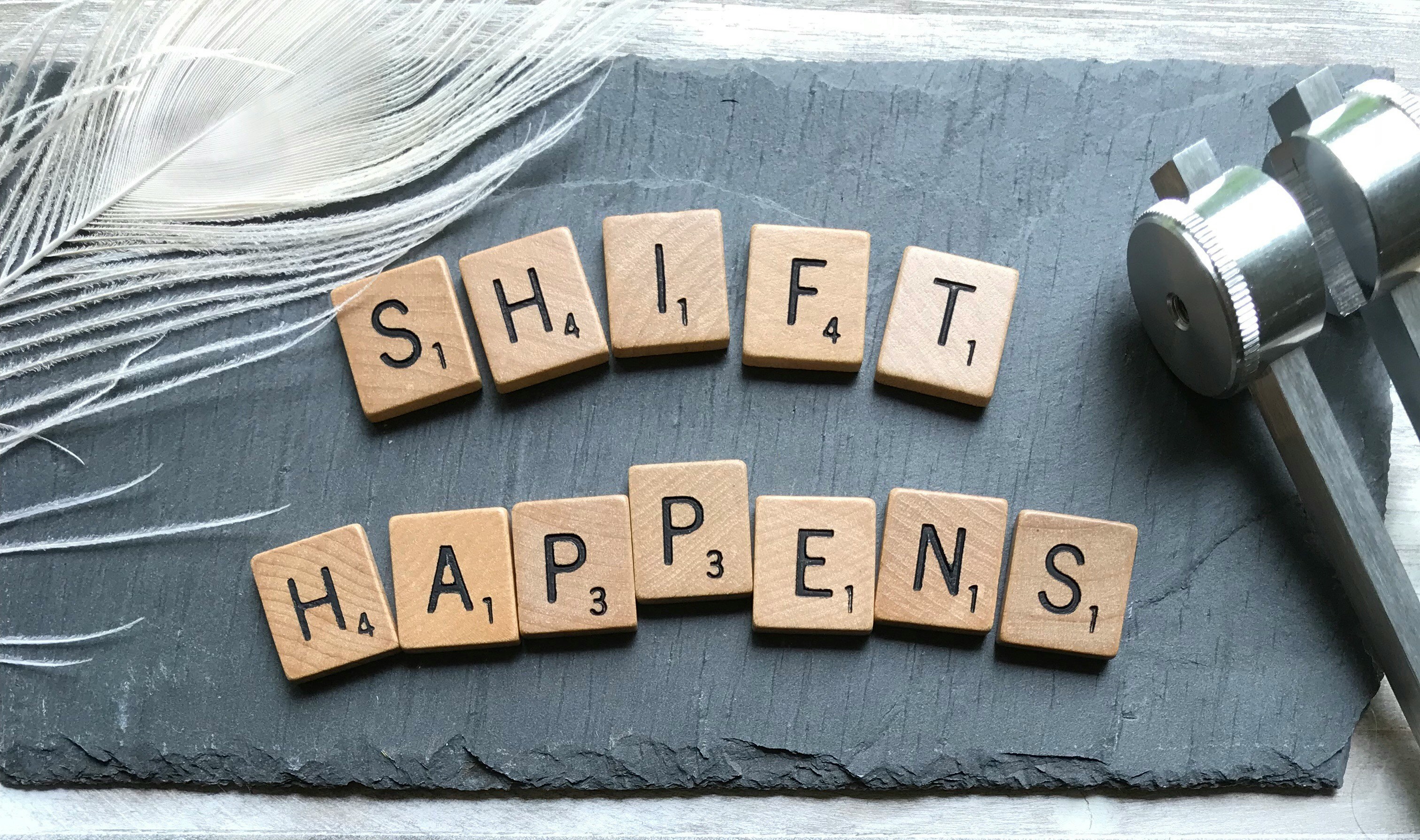 Scrabble tiles spelling the phrase “SHIFT HAPPENS” arranged on a slate surface beside a white feather and a metal caliper, symbolizing change and transformation.