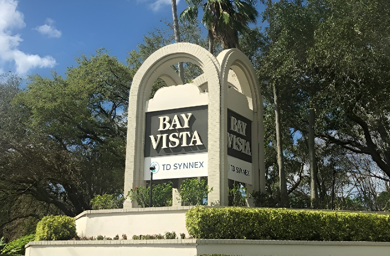 A large outdoor sign for Bay Vista with a TD Synnex logo sits on a landscaped concrete base surrounded by trimmed hedges, tall trees and bright greenery under a blue sky.