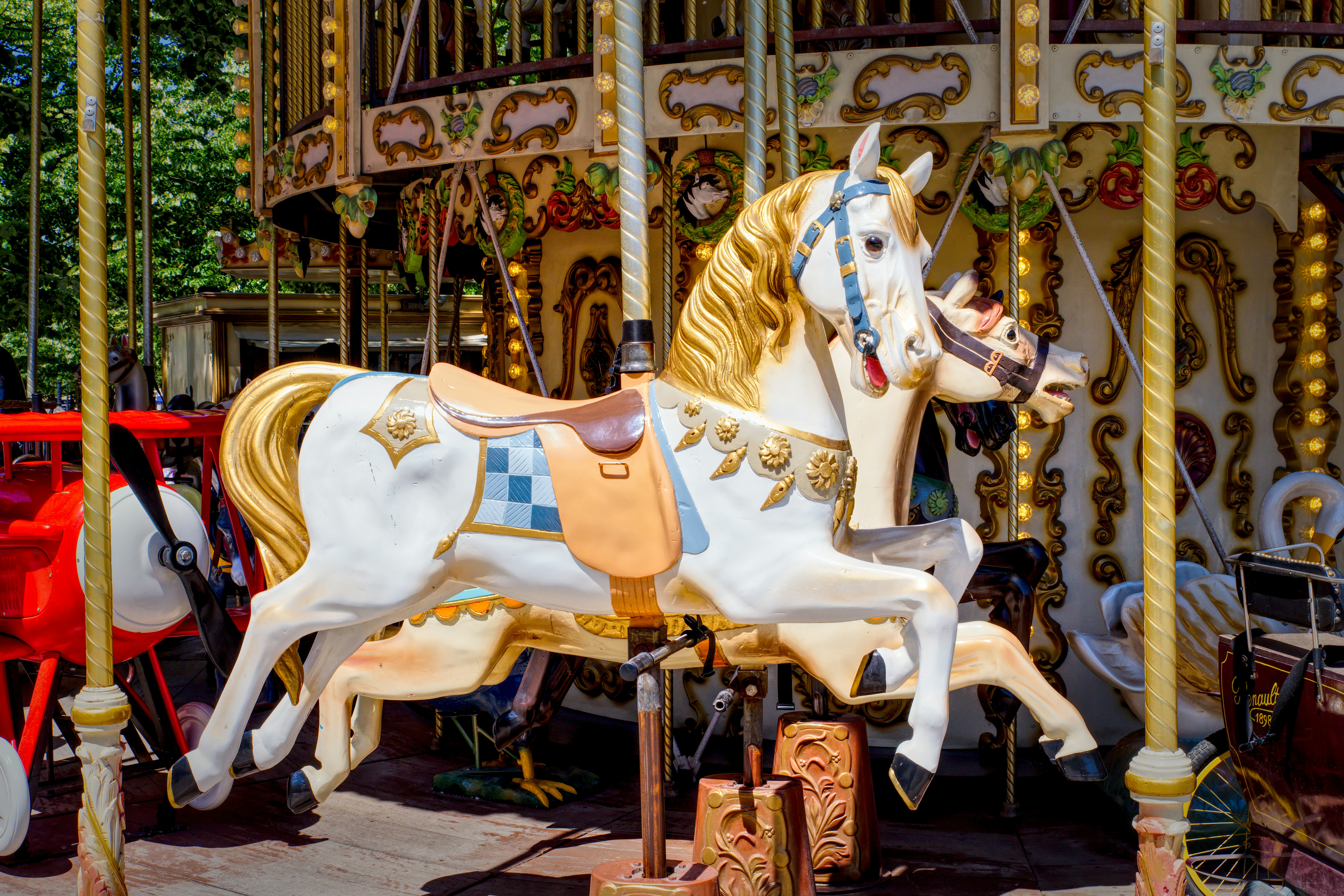 Close-up of a decorative white carousel horse with a golden mane and ornate saddle, set against the colorful, detailed backdrop of a traditional merry-go-round.