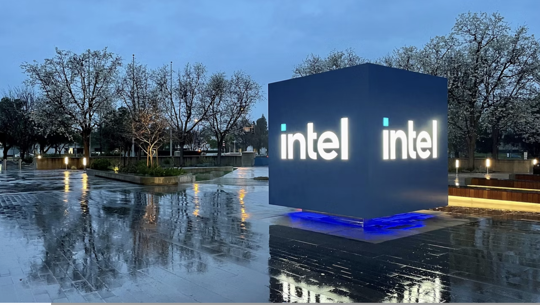 Illuminated Intel logo on a cube installation at an outdoor campus plaza with wet pavement.