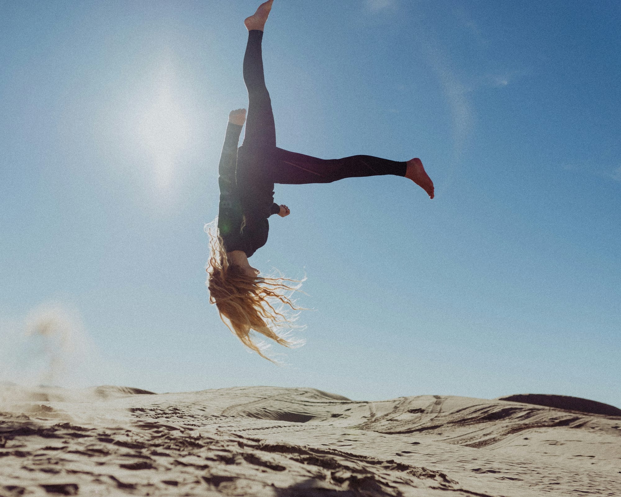 Person mid-flip above sandy dunes under a clear blue sky.