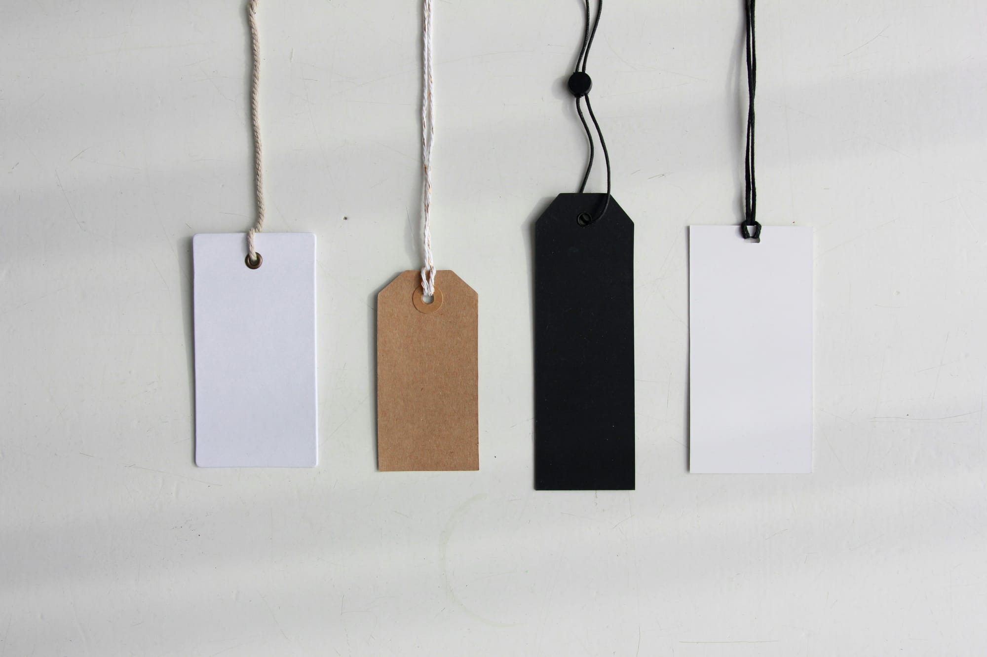 Four blank hanging price tags in white, brown and black attached to strings against a light background.