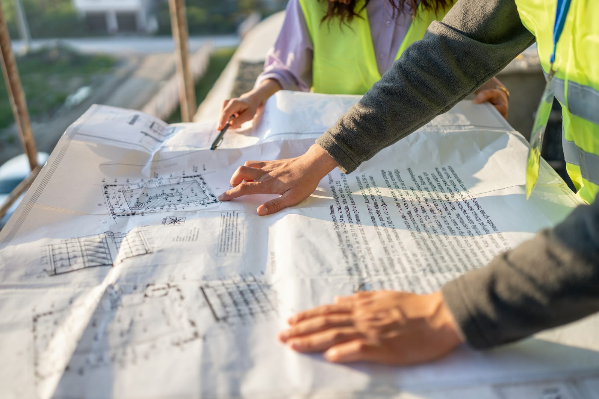 Two people in high-visibility vests review architectural blueprints on a construction site.